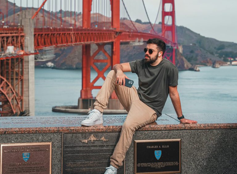 Man with glasses on sitting on wall. Behind him is the Golden Gate Bridge