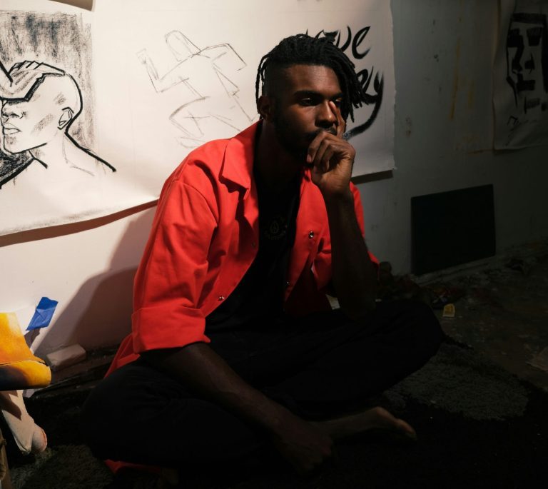 A young male artist squats with his hand on his chin with art in the background.